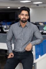 Portrait of handsome young Indian businessman casually standing against his workstation. Corporate office environment.