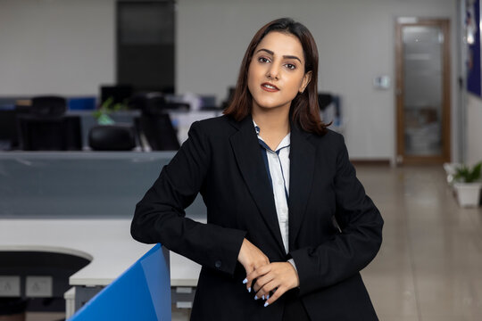 Portrait of young successful Indian business woman in black suit standing against office background.