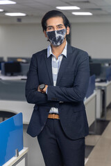 Portrait of handsome young Indian businessman wearing Covid protection mask, standing against office background. Corporate office environment.