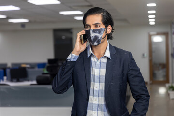 Portrait of handsome young Indian businessman wearing Covid protection mask, speaking on his mobile phone, Corporate office environment.