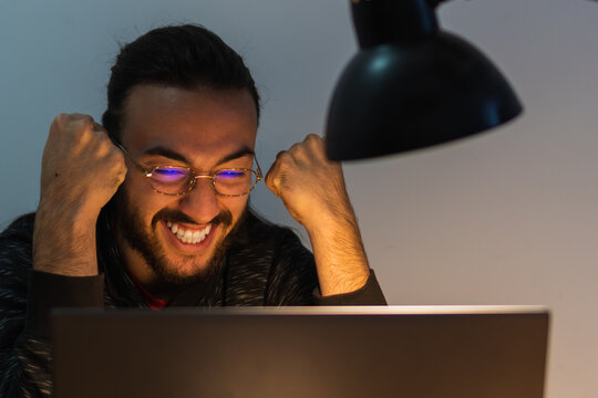 Animated Latino Student Looking At Test Results On A Laptop Computer, Wearing Glasses. Fists In Hands, Smiling.