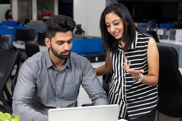 Portrait of two young smiling businesspeople looking at laptop and discussing project at workstation, corporate environment.