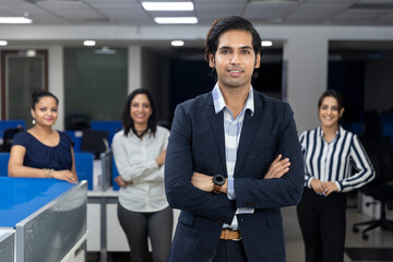 Confident Indian businessman standing infront of his office colleagues, selective focus, corporate environment, team members, business office.