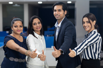 Four Indian office colleagues standing together against office background, showing thumbs up sign of victory, corporate environment.