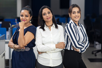 Three Indian businesswomen with arms folded standing together against office background, corporate environment.