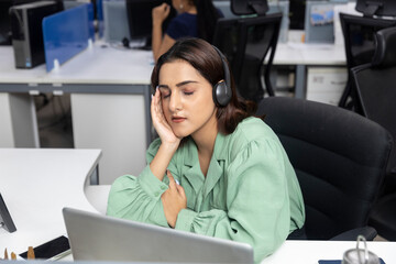 Portrait of sleepy Indian businesswoman wearing headphones, resting face in her palm, corporate environment, call centre employee.