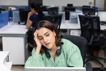 Portrait of sleepy Indian businesswoman wearing headphones, resting face in her palm  corporate environment, call centre employee.