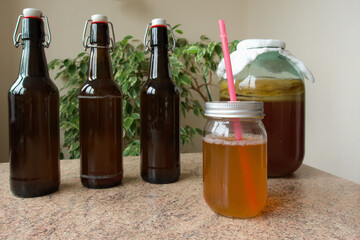 Large jar containing tea and tea mushroom, covered by cheesecloth, along with three bottles of ready to drink kombucha and a mason  jar with the beverage