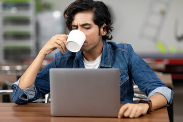 Portrait of a young handsome Indian man sipping from a coffee mug, sitting in an office cafeteria, coffee shop, casual work environment.