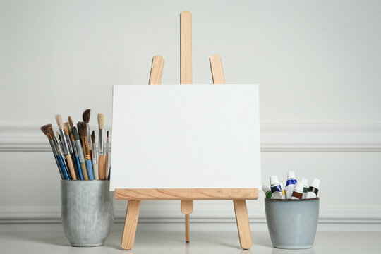 Easel with blank canvas, paints and brushes on white wooden table