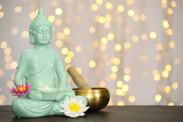 Buddha statue with candle, lotus flowers and singing bowl on table against blurred lights. Space for text