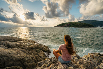 Young woman meditating at the seaside sitting in the lotus position on rocks overlooking the sea with sunset in a healthy lifestyle concept