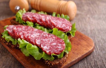 Delicious sandwich on black grain bread with lettuce and smoked sausage on old wooden background.