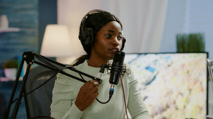 African woman host of online show talking into microphone wearing headphones. Speaking during...