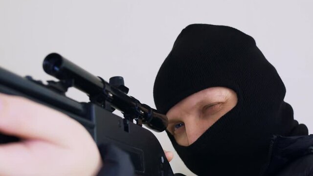 A terrorist in a black balaclava raises a sniper rifle, watches the target. Contract murder. Close-up.