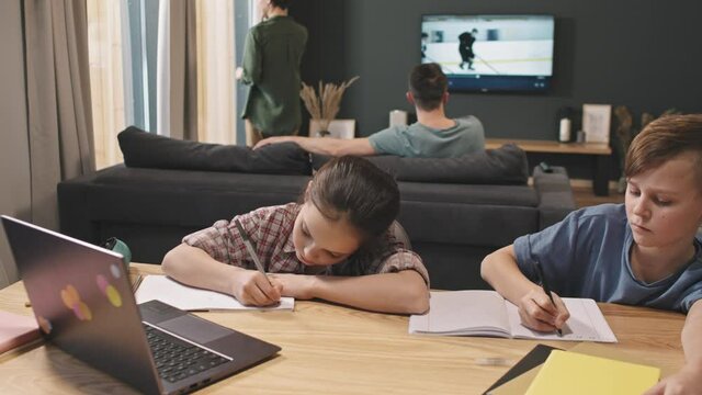 PAN waist-up shot of cute elementary age brother and sister studying online on laptop writing in their copybooks while their mom talking on phone and dad watching hockey match on tv in background