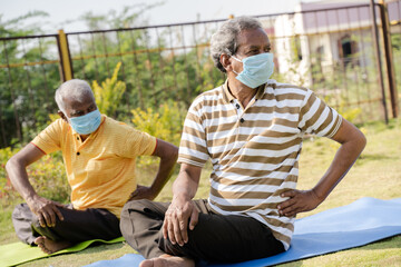 Two active senior people with medical face mask exercising on yoga mat during early morning - concept of new normal, elderly helthcare and fitness class at park during coronavirus covid-19 pandemic