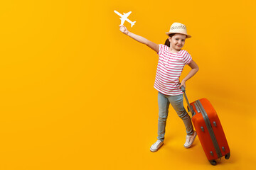 Happy little preteen girl holding a paper airplane and dreaming about travel. Child with a suitcase on a yellow background with copy space for text.