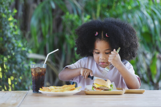 childhood and eating concept - little african american curly hair girl used knife slice burger and french fries on the table .enjoying unhealthy food. - Powered by Adobe