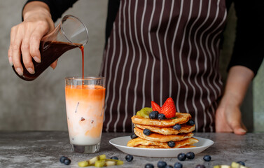 Hand of woman housewife in apron pouring fresh tea from bottle in to milk glass with ice beside stack of homemade pancakes with fruit topping