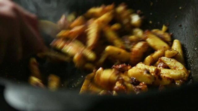 The cook's hand with a slotted spoon is stirring a pieces of potatoes are fried in oil in a cast-iron frying pan with a round bottom, steam comes, the oil boils. Cooking potatoes in cauldron, close-up