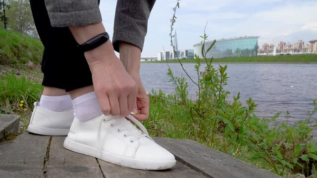A man ties his shoelaces on white sneakers on the street against the background of the river and nature close-up.