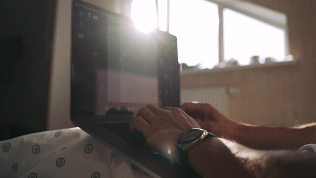 Men's hands are typing on a laptop Beautiful rays of the sun fall through the window The shadow of the sunset lies on the bed where the employee is working on the computer Slow motion