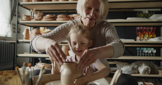 Grandmother teaches her granddaughter working on a pottery rotating wheel. Senior woman and cute little girl make pot on a spinning pottery wheel. Hobby, leisure, arts and crafts concept.