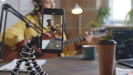 Selective focus shot of smartphone on tripod filming female musician playing the guitar and singing into microphone in recording studio during live stream - Powered by Adobe