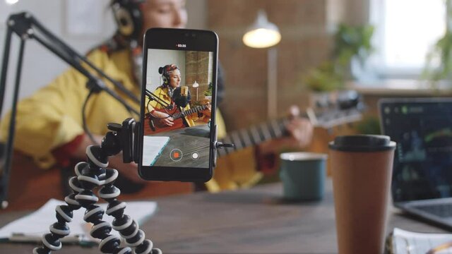 Selective focus shot of smartphone on tripod filming female musician playing the guitar and singing into microphone in recording studio during live stream