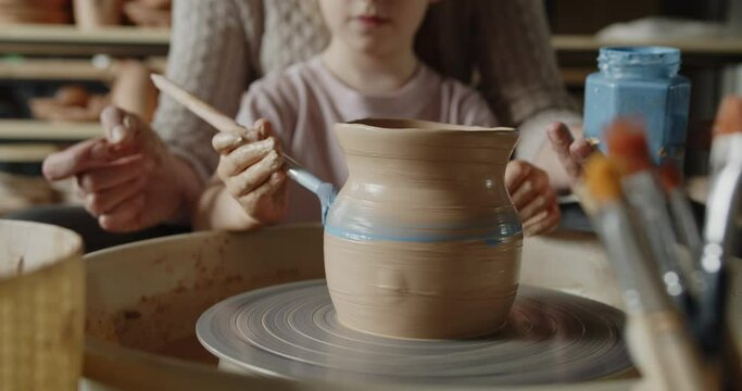 Grandmother teaches her granddaughter working on a pottery rotating wheel. Senior woman and cute little girl painting handmade pot on a spinning pottery wheel. Hobby, leisure, arts and crafts concept.