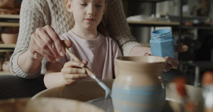 Grandmother teaches her granddaughter working on a pottery rotating wheel. Senior woman and cute little girl painting handmade pot on a spinning pottery wheel. Hobby, leisure, arts and crafts concept.