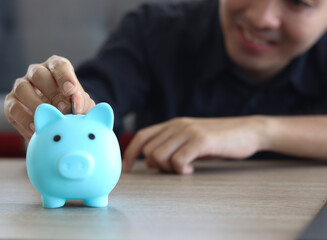 Obraz na płótnie Canvas Happy smiling young Asian man collecting coins in blue piggy bank on desk, finance savings or investment for the future concept.