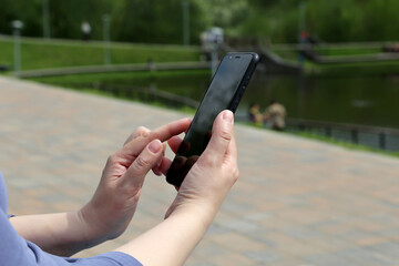 Woman with smartphone sitting in summer park. Close up of female hands with mobile phone on green nature background, concept of sms, online addiction, social media
