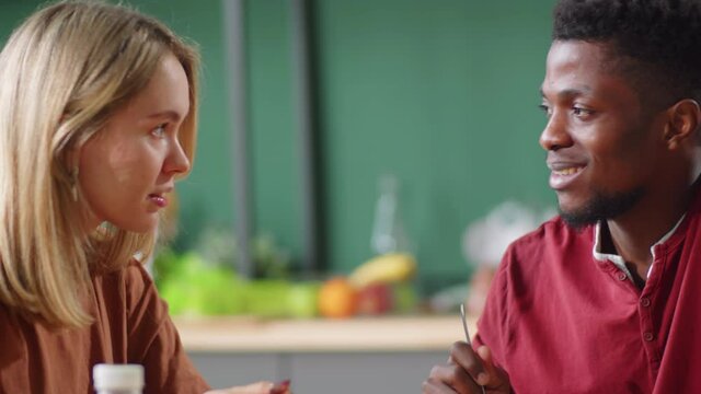 Young black man and caucasian woman sitting together at kitchen table, eating healthy food from ecofriendly containers and chatting over meal