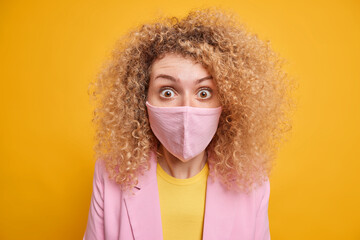 Close up shot of curly haired woman wears protective mask shocked by lockdown restrictions and quarantine extend faces risk of infection spreading dressed in formal clothes isolated over yellow wall