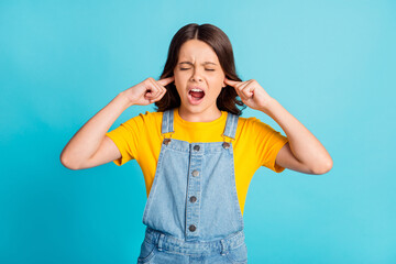 Photo of annoyed irritated small school girl wear denim jeans overall fingers close ears screaming isolated blue color background