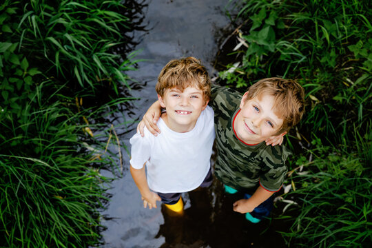 Two happy little school kids boys, funny siblings having fun together walking through water in river in gum rubber boots. Family portrait of healthy brothers and best friends
