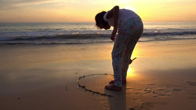 Beautiful girl writing heart on sand at sunset or sunrise Amazing beautiful light of nature on sandy shore Sea surface