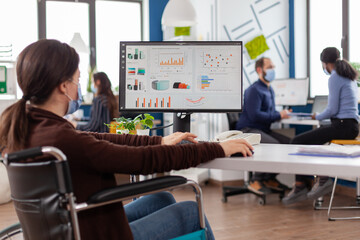 Immobilized business woman with protective mask working in new normal business financial company typing on pc, checking reports, analysing data looking at desktop sitting in wheelchair.