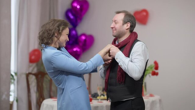 Side view portrait of loving happy adult man kissing hands of beautiful woman talking compliments to partner. Smiling Caucasian husband enjoying Valentine's anniversary celebration with wife indoors