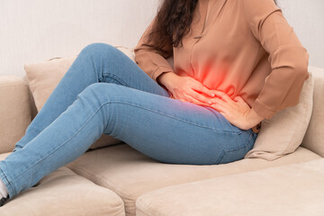 Unhappy Asian woman sitting on the sofa and holding on stomach suffering. Abdominal pain that comes from menstruation, diarrhea, or indigestion. Sickness and healthcare concept - obrazy, fototapety, plakaty