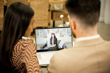 Rear view of young married couple wife and husband discussing issues and misunderstandings in relationships when having online consultation with family psychologist, sitting on sofa in livinng room