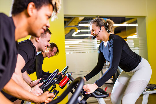 Pretty Authentic Female Instructor With Headset In Fitness Class Exercise With Group In Cycling Room