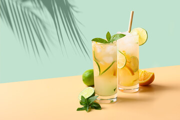 Two summer cold mojito cocktails with orange and lime slices on green and beige background. Refreshing summer beverage with sunny shadow. - obrazy, fototapety, plakaty