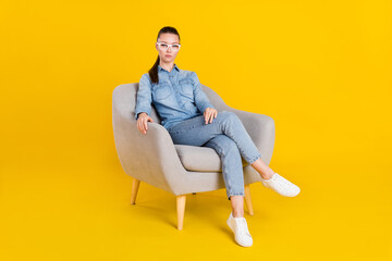 Photo of serious confident young woman wear denim shirt glasses sitting chair isolated yellow color...