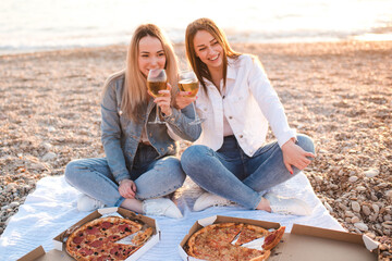 Two young beautiful girlfriends having fun with pizza and wine at beach over sea shore outdoors in sun light. Summer vacation season. Friendship. Happiness. Sisters spend time together at coast line.