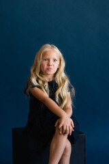 beautiful blonde in a dark blue dress on a solid blue background. The photo is great for demonstrating the character of a child - daydreaming, enthusiasm, vulnerability, touching, beauty, stubbornness