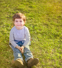 A small cute child in jeans sits on the grass in the rays of the setting sun and smiles. Summer scene