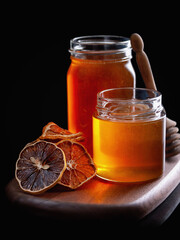 Honey dipper stands behind jars of honey standing on a wooden tray next to dried oranges and lemons on a black background